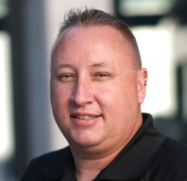 A smiling man with short hair wearing a black polo shirt. The background is softly blurred, suggesting an outdoor or urban setting.