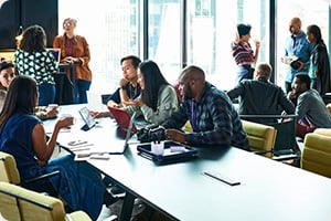 A diverse group of people engage in discussion and collaboration around a large table in a modern office with large windows, conveying a lively, busy atmosphere.