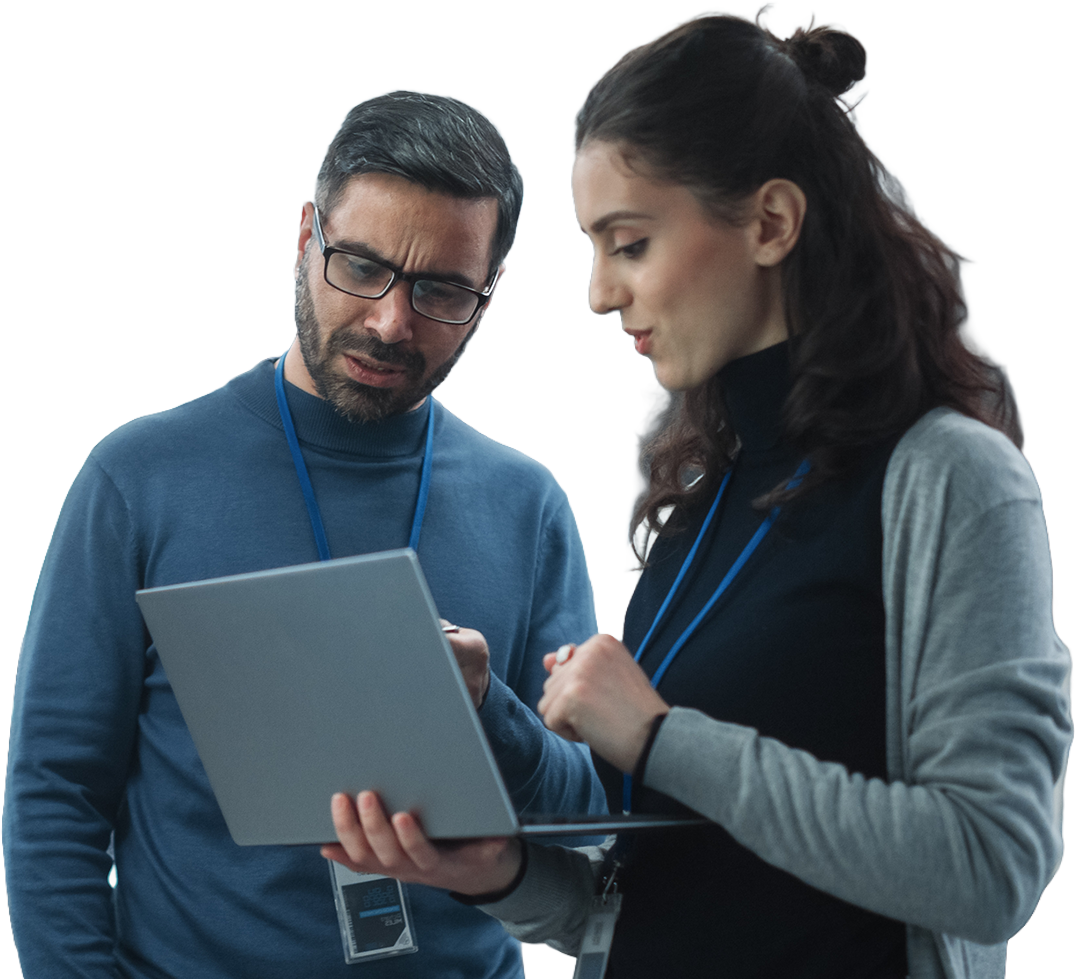Two individuals wearing lanyards, engaged in discussion while looking at a laptop. The scene conveys focus and collaboration against a dark background.